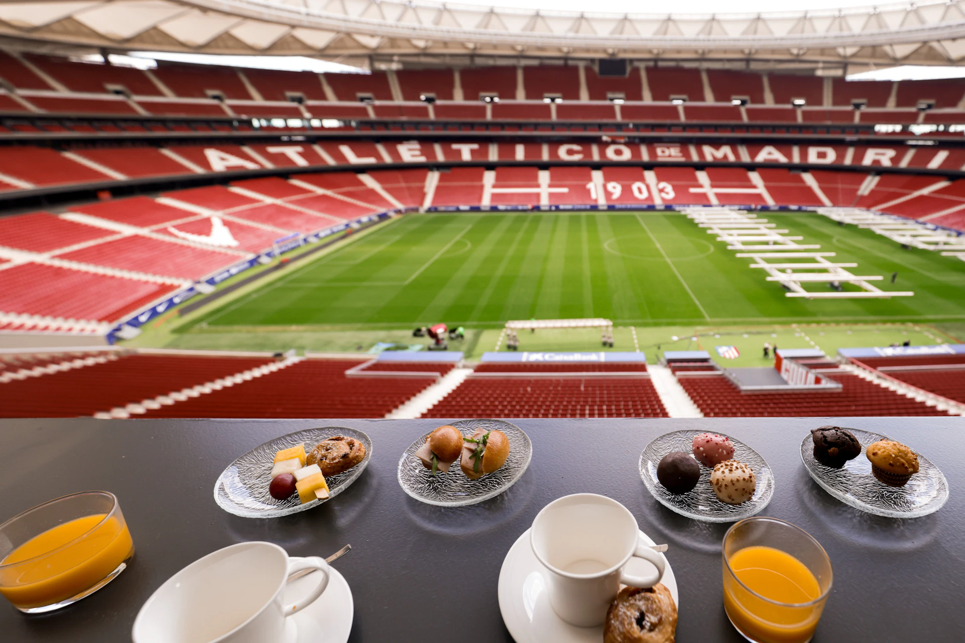 A view of a soccer stadium with a green field, seen from a suite with small plates of snacks and cups on a table.