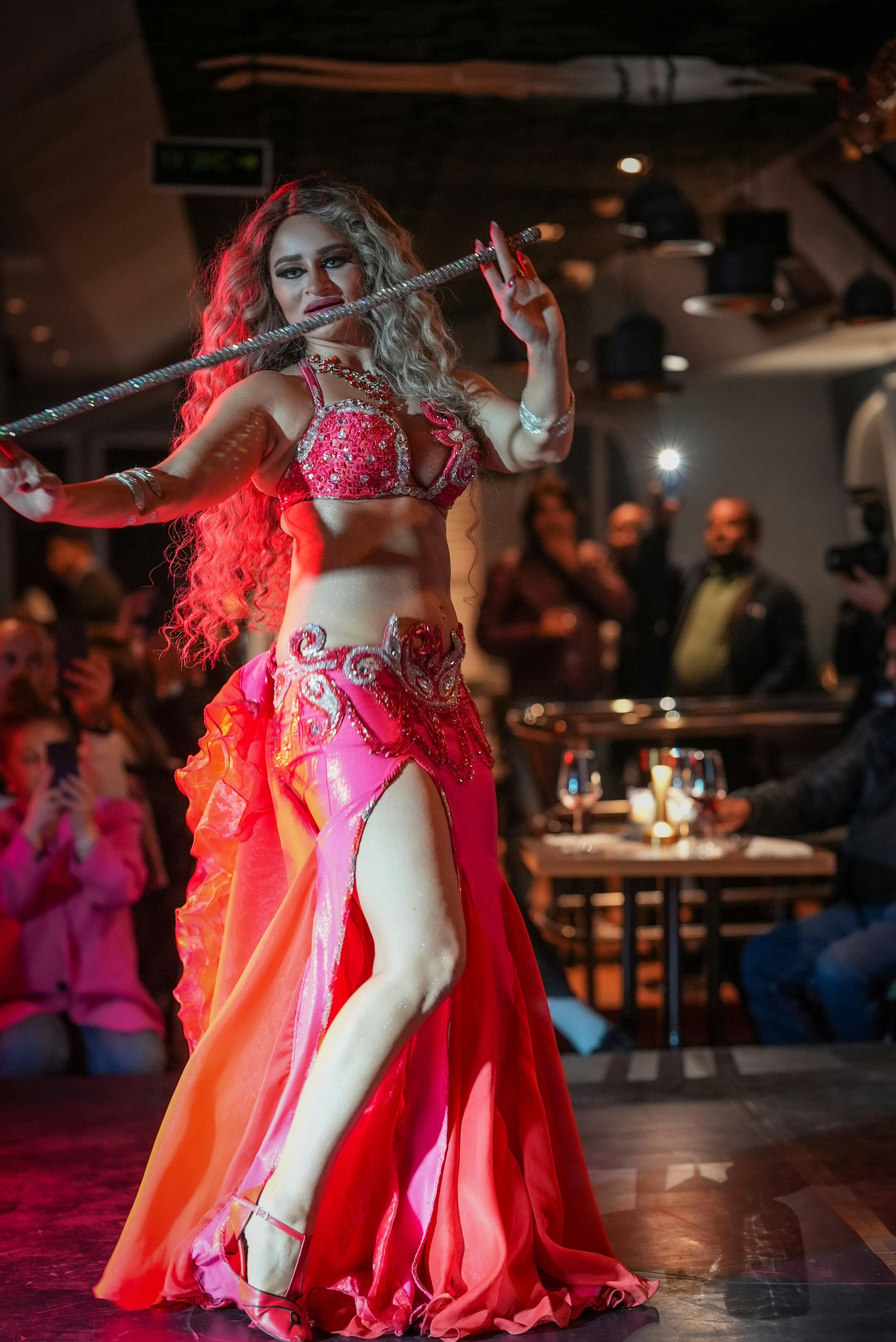 明るいピンクのコスチュームを着た女性が、見物人を背景に、薄暗いレストランで剣を持ってダンスを踊っています。