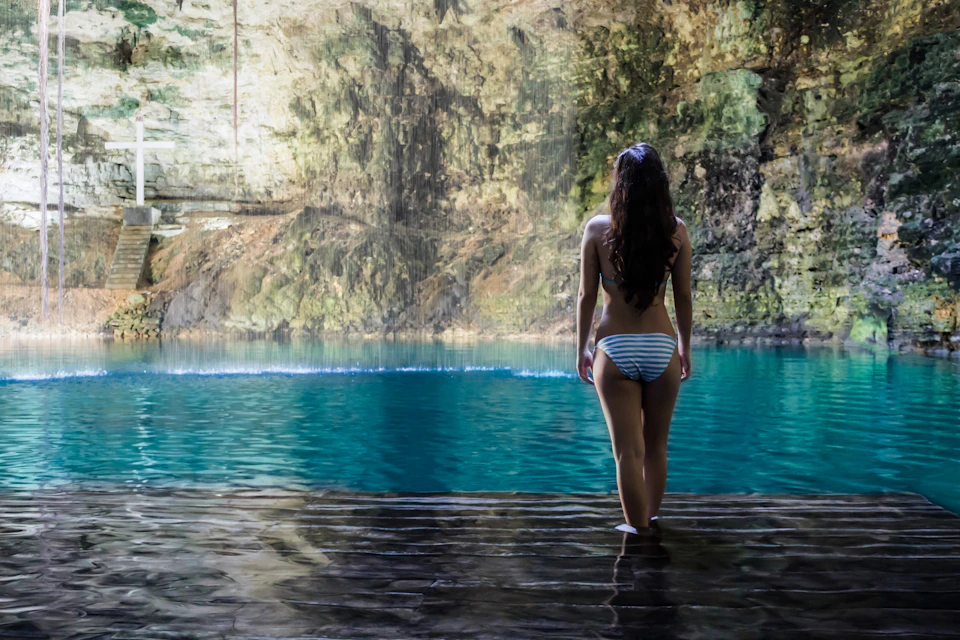 A woman stands in front of a blue water pool inside a cave, with sunlight illuminating the rocky walls.