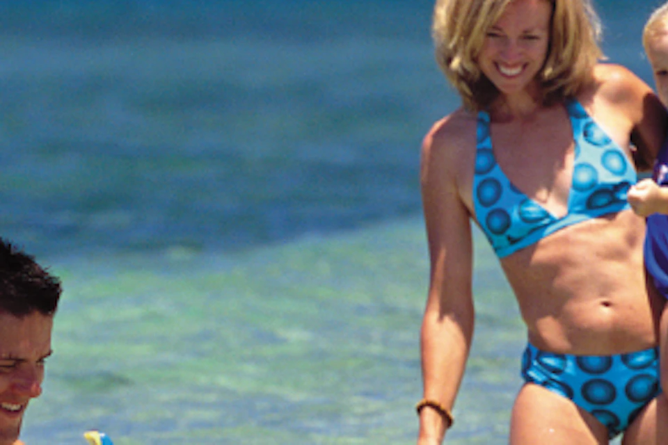 A man and woman with two children enjoy the shallow, clear water at a beach. One child snorkels while the other is held by the woman.