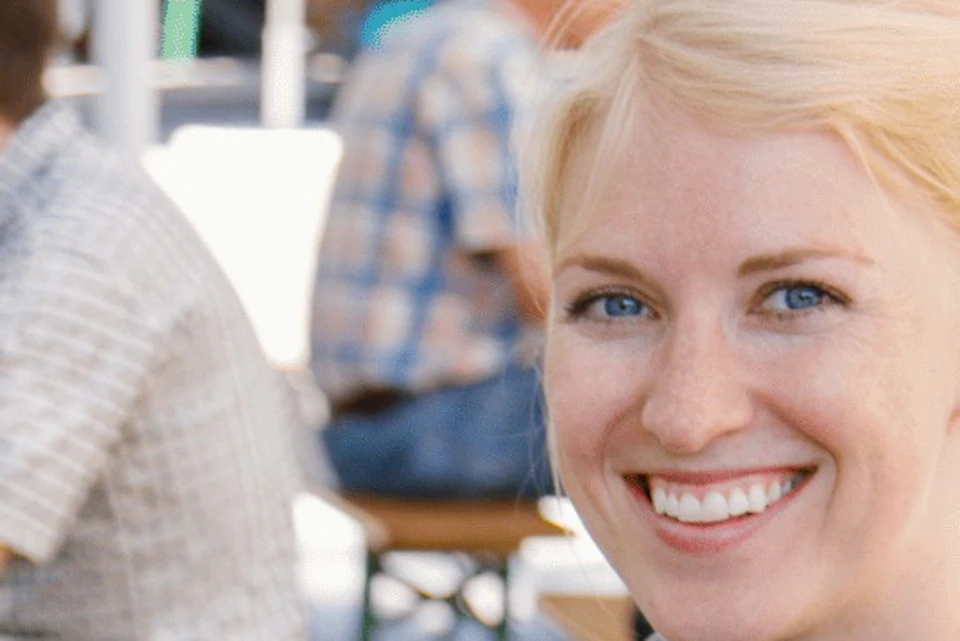 A smiling woman holding two beer glasses, sitting outdoors at a table. Other people are seen seated in the background.