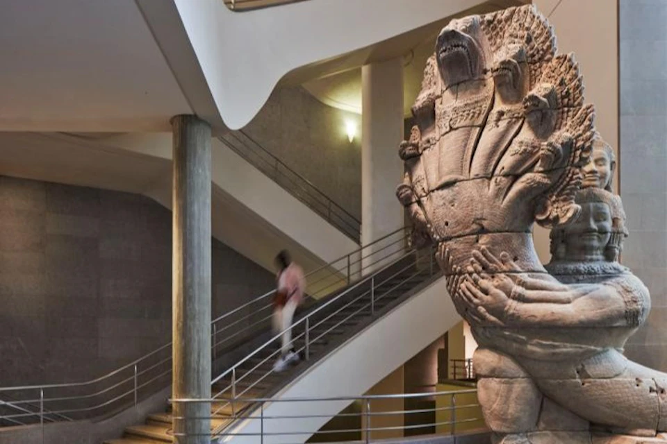 A large stone sculpture of a multi-headed serpent with a human figure in a museum setting, with a staircase in the background.