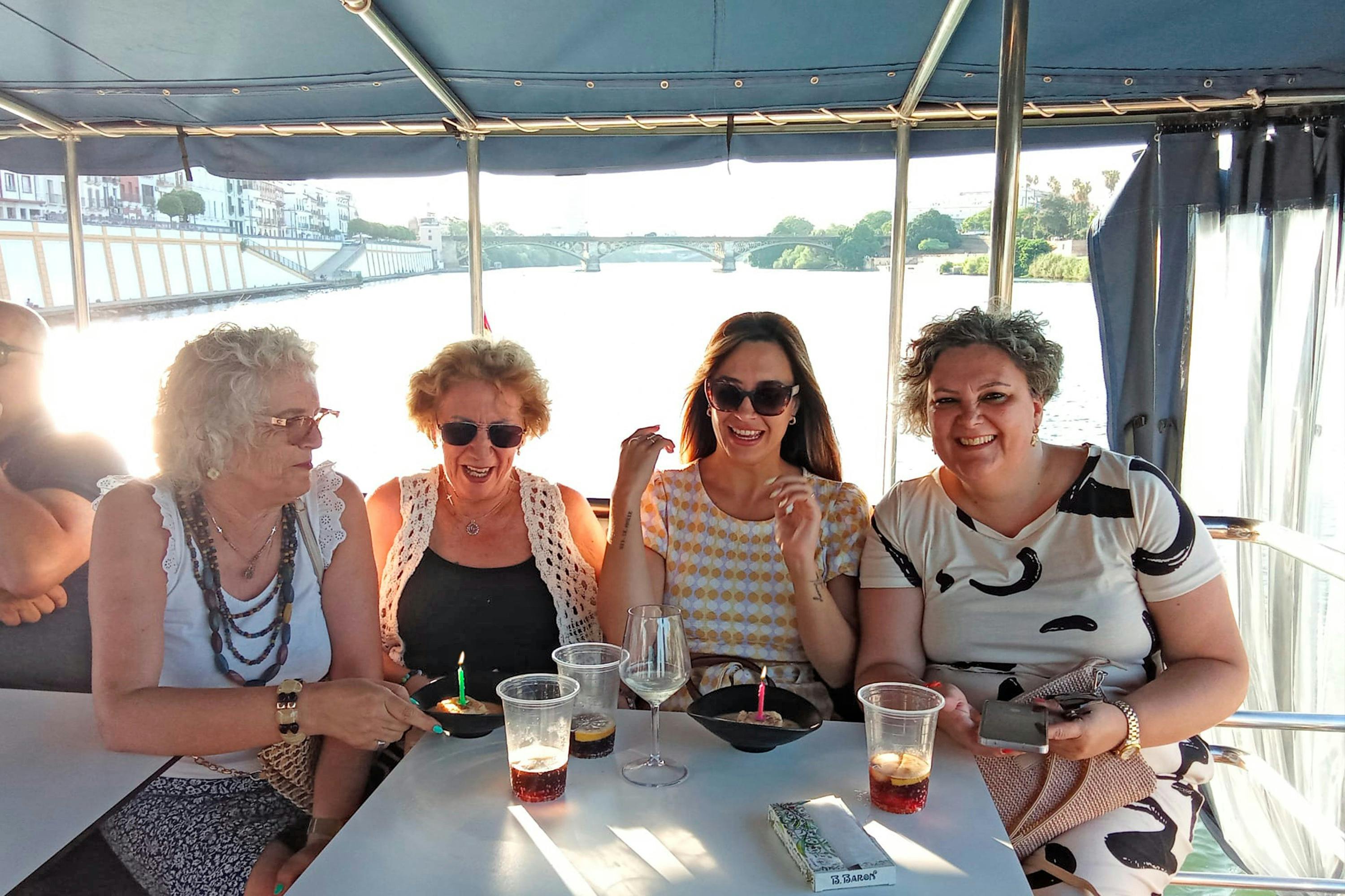 Three women seated at a table on a boat, smiling and enjoying drinks, with a river and a bridge in the background.
