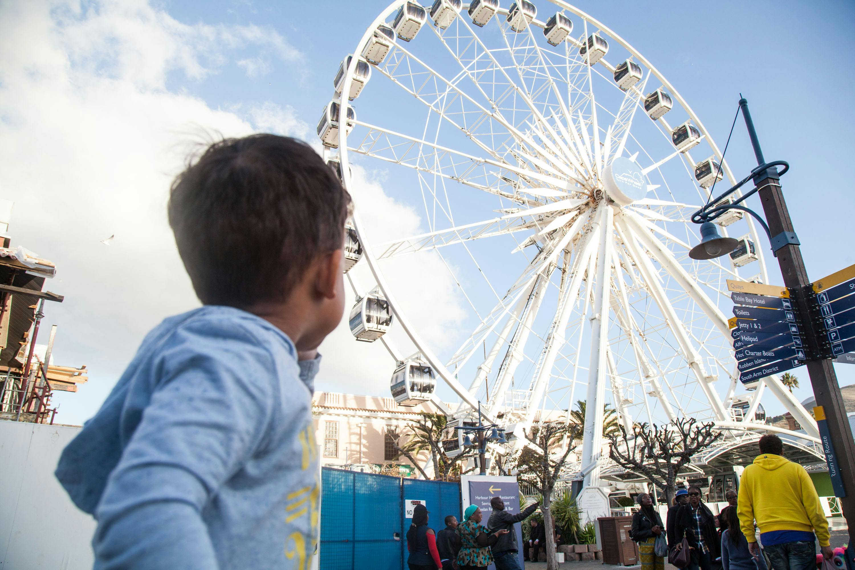 화창한 날 한 어린이가 커다란 대관람차를 바라보고 있는데, 기지 근처에 사람들이 서 있고 방향 표지판이 보입니다.
