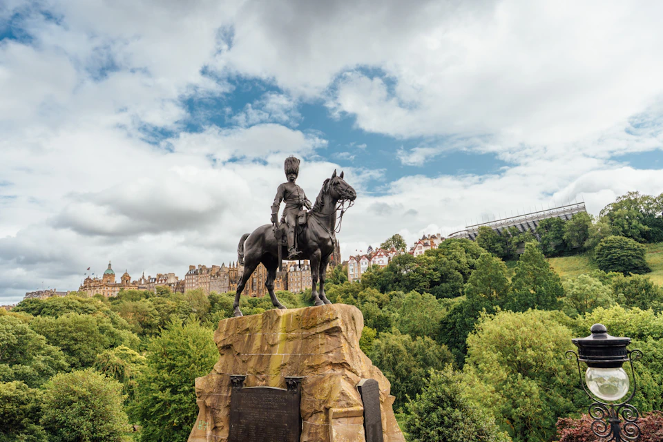 Equestrian statue on a stone pedestal in a park setting with trees, buildings, and a cloudy sky in the background.
