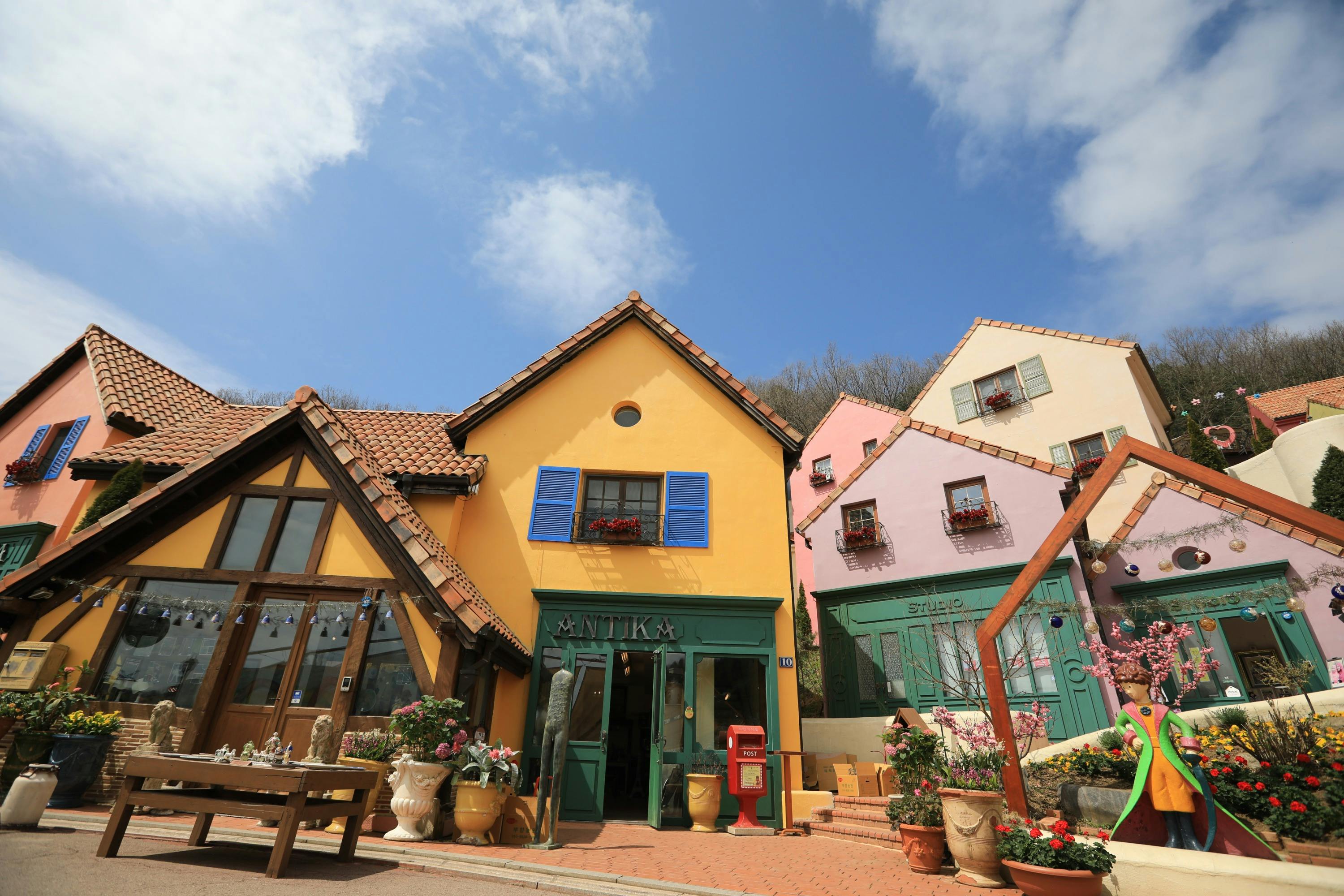 A colorful village with yellow and pink buildings, blue shutters, and decorative plants under a blue sky with clouds.