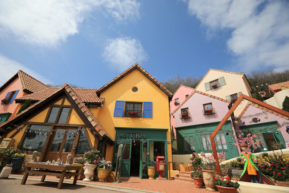 A colorful village with yellow and pink buildings, blue shutters, and decorative plants under a blue sky with clouds.