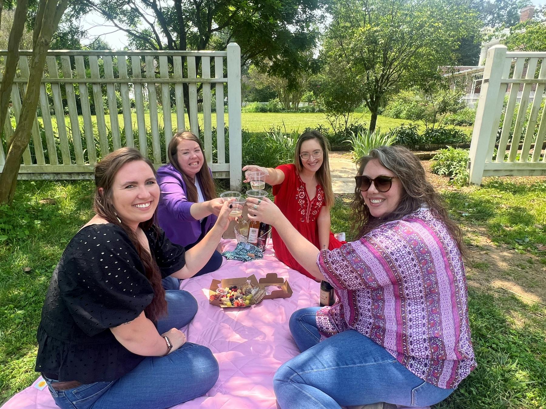Four women having a picnic on a pink blanket, toasting drinks, with food boxes in front, near a white fence and trees.