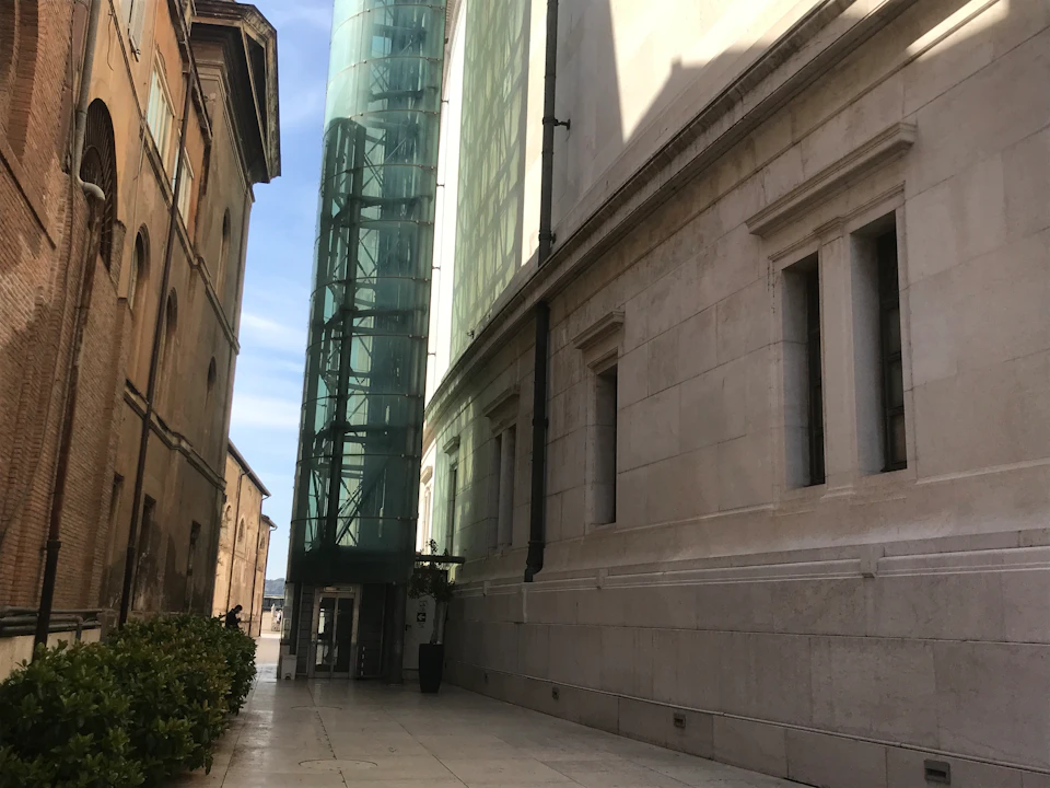 Narrow alley between buildings with a glass elevator shaft on the left, windows on the right, and bushes in the distance.