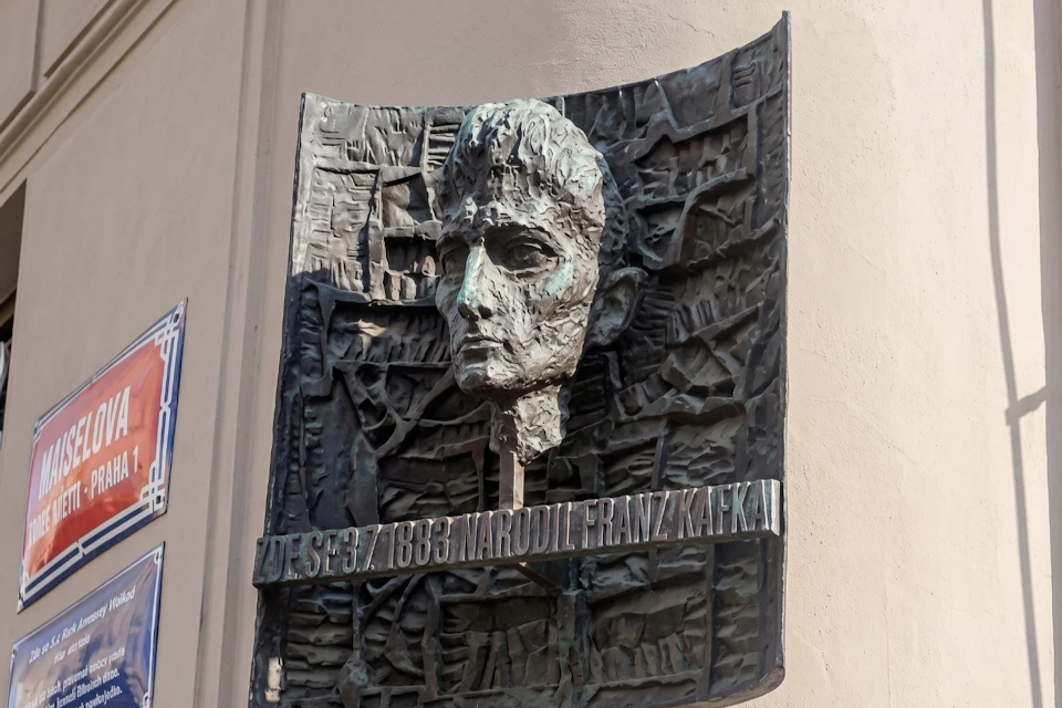 A bronze relief of a face with text stating "Zde se 3.7.1883 narodil Franz Kafka" mounted on a wall.