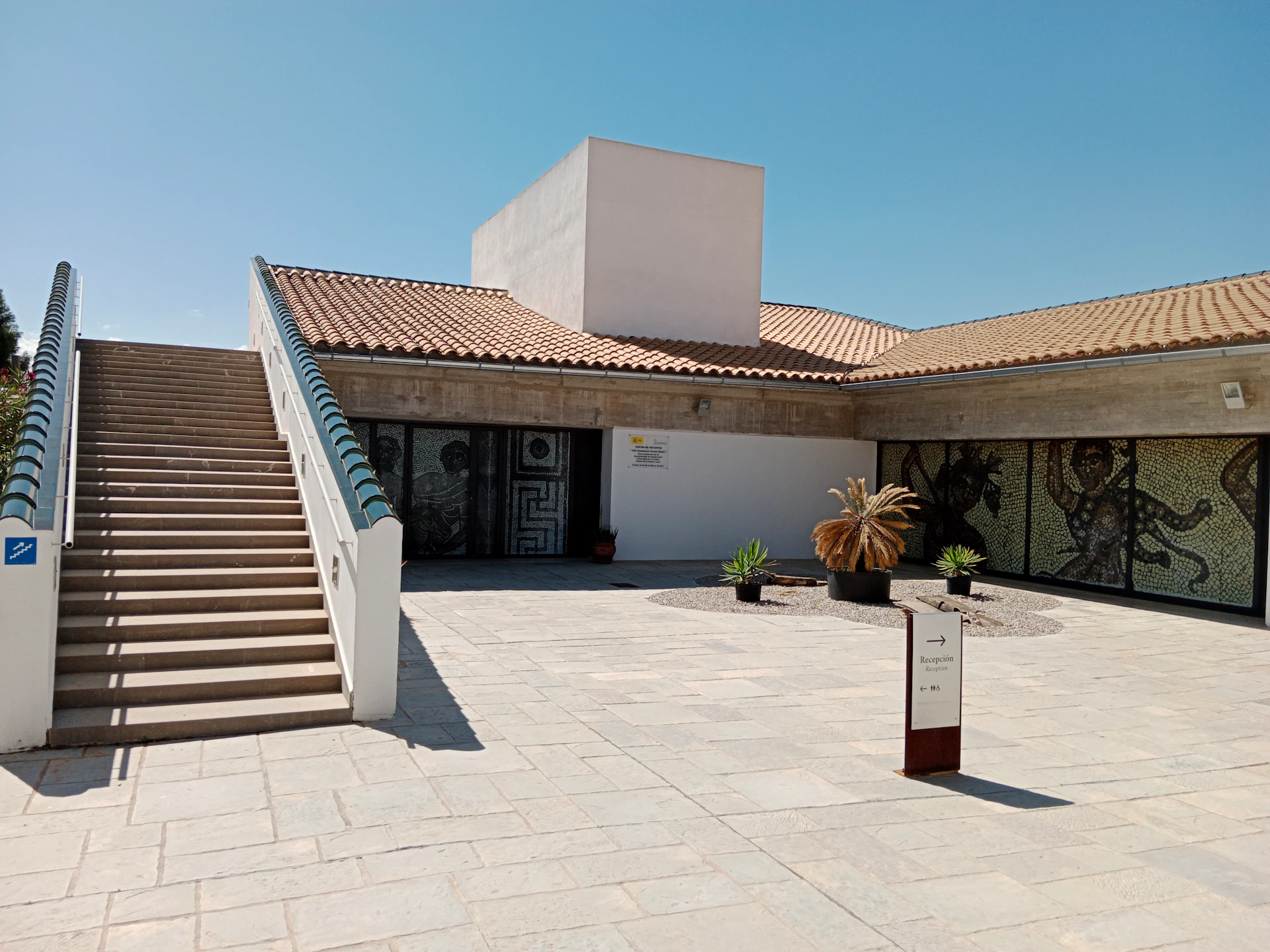 A courtyard with tiled ground, stairs, potted plants, a modern building with tiled roofs, and a signpost under a clear sky.