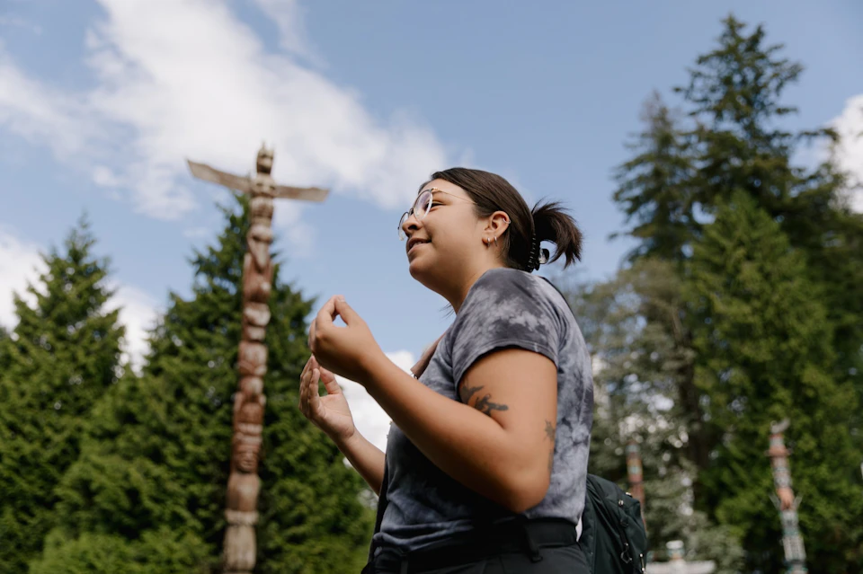 A person outdoors wearing casual clothing and glasses, gesturing with their hands, with a totem pole and trees in the background.