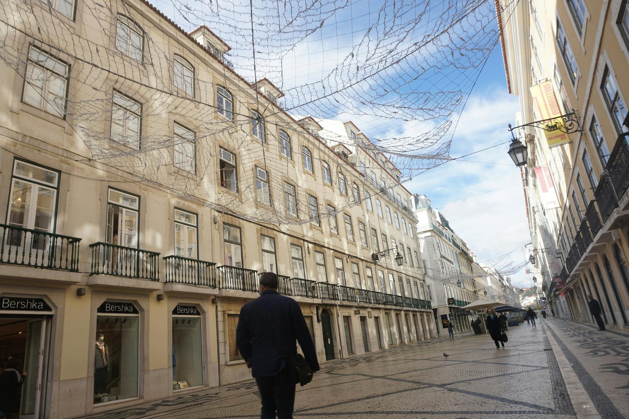 A person walks down a narrow, cobblestone street lined with buildings. Overhead, decorative netting spans between buildings.