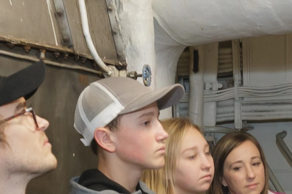 A group of people stands inside a mechanical room while a guide gestures towards pipes and equipment.