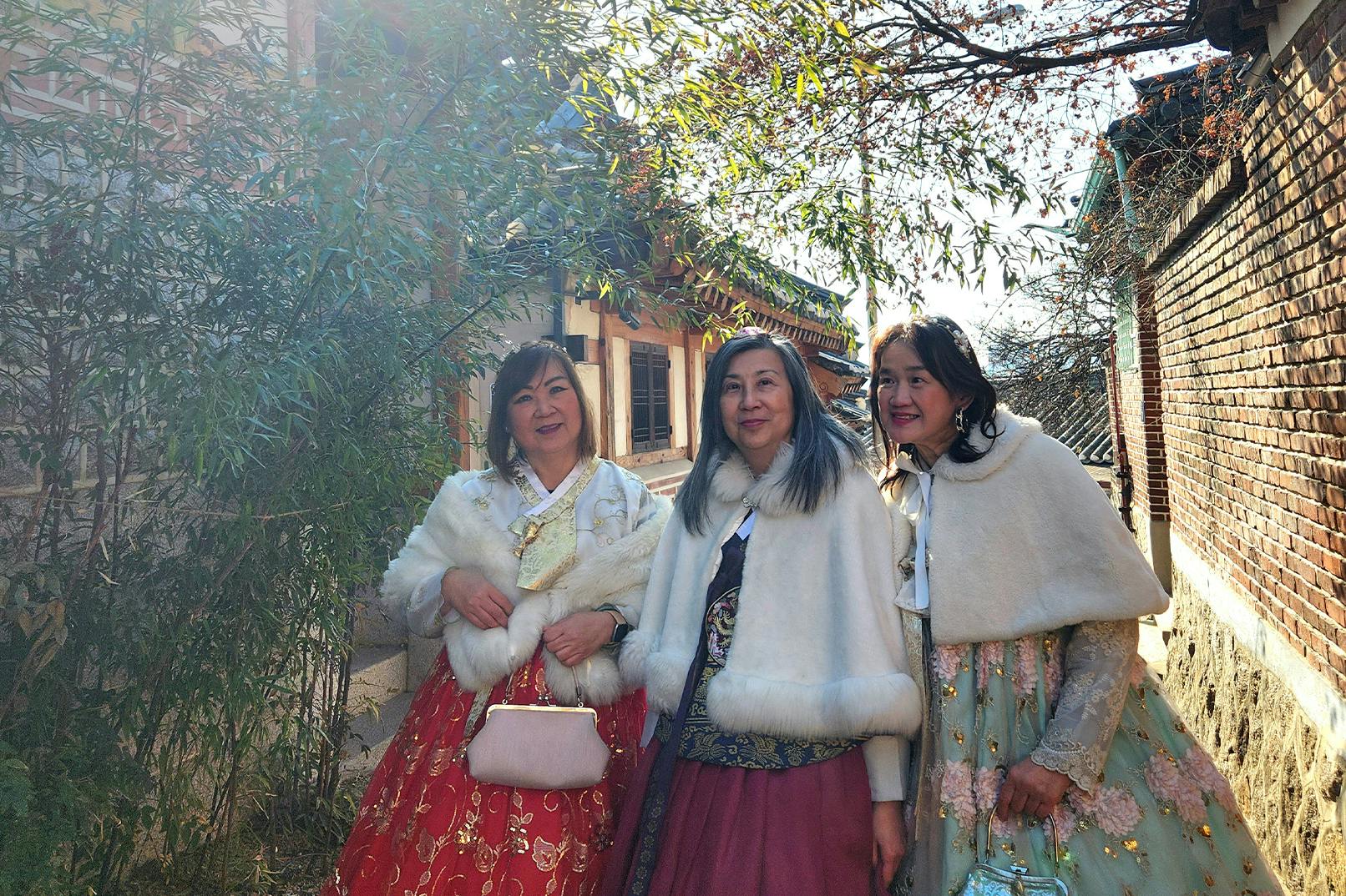 Trzy kobiety w tradycyjnym stroju pozują razem na świeżym powietrzu pod ścianą i roślinami bambusa, w świetle słonecznym.