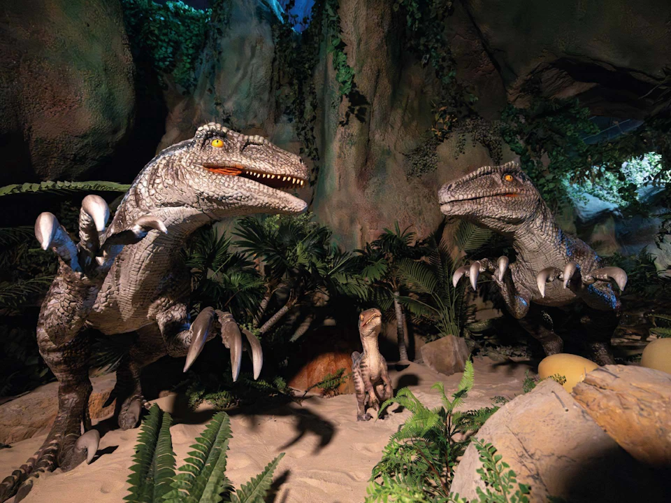 A display of dinosaur models in a jungle-like setting, featuring two large dinosaurs and a baby dinosaur among vegetation.