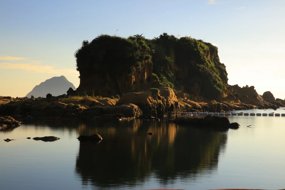 A small, rocky island covered in greenery, surrounded by calm water with a mountain visible in the background.