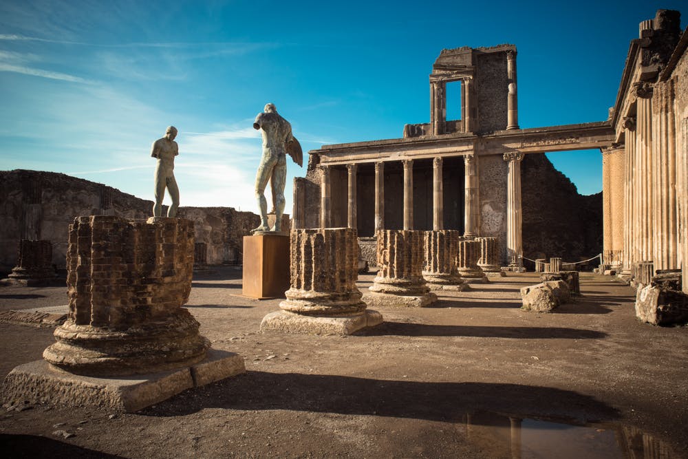 Ancient ruins with stone columns, two statues, and a partially intact rectangular building under a clear blue sky.