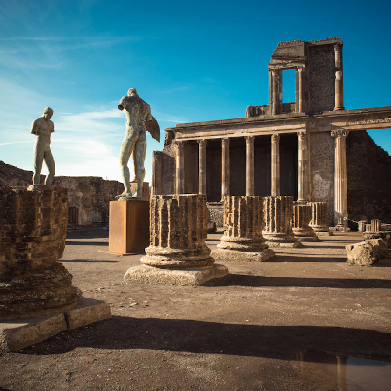 Ancient ruins with stone columns, two statues, and a partially intact rectangular building under a clear blue sky.