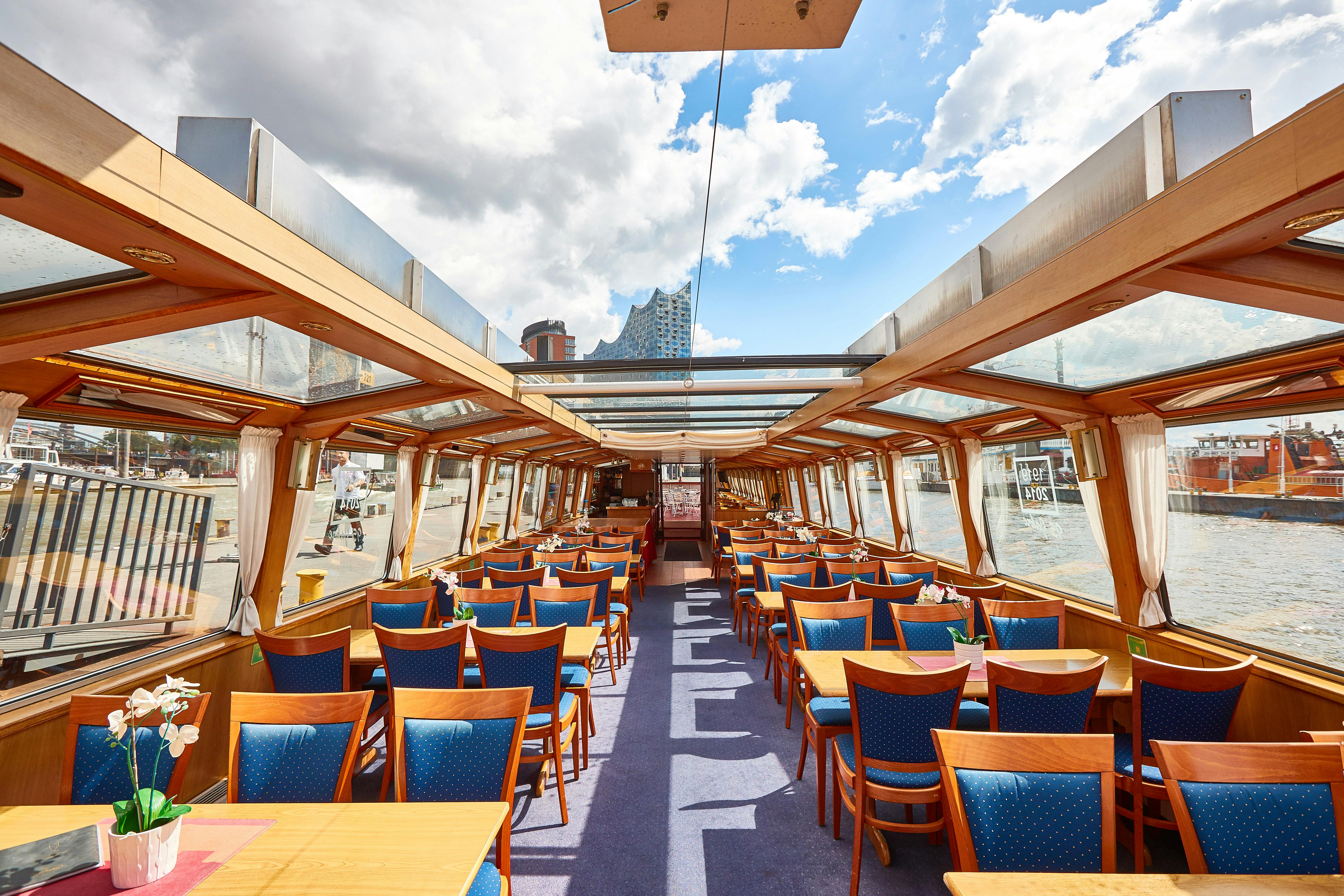 En tom spiseplads inde i en båd med træborde og blå polstrede stole, store vinduer og et glasloft under en delvist overskyet himmel.