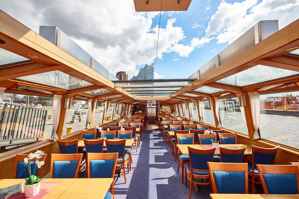 An empty dining area inside a boat with wooden tables and blue cushioned chairs, large windows, and a glass ceiling under a partly cloudy sky.
