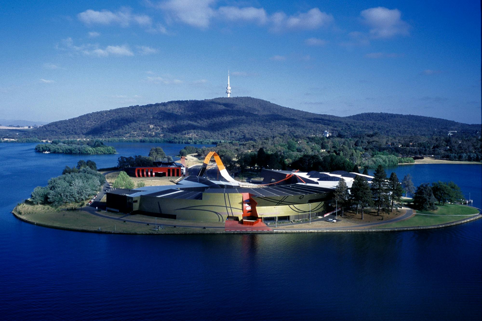 Vue aérienne d'un bâtiment moderne à l'architecture colorée sur une péninsule entourée d'un lac et de collines en arrière-plan.