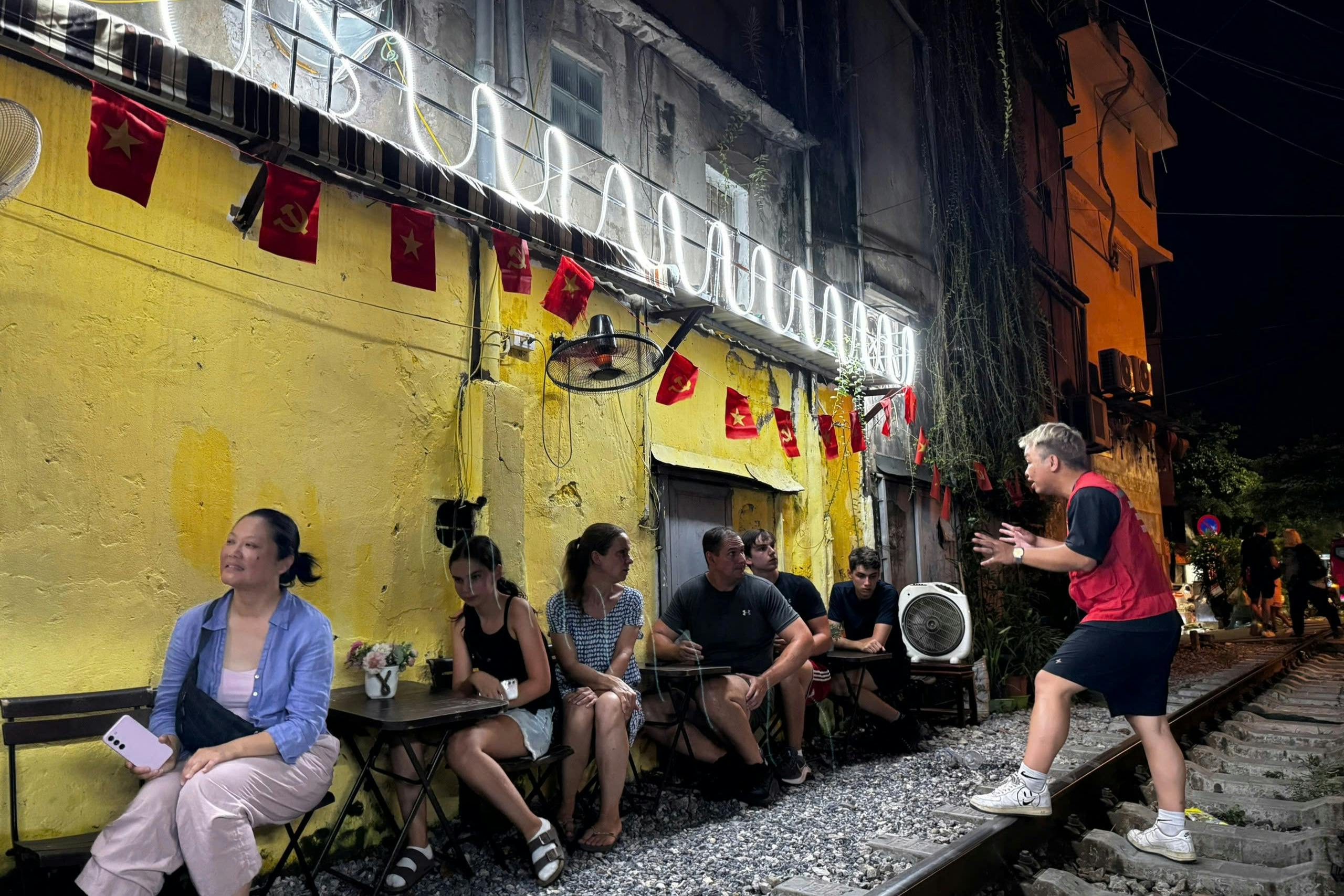 Le persone sono sedute su sedie con tavolini lungo un edificio giallo ornato di bandiere, mentre un'altra persona parla animatamente, in piedi sui binari della ferrovia.