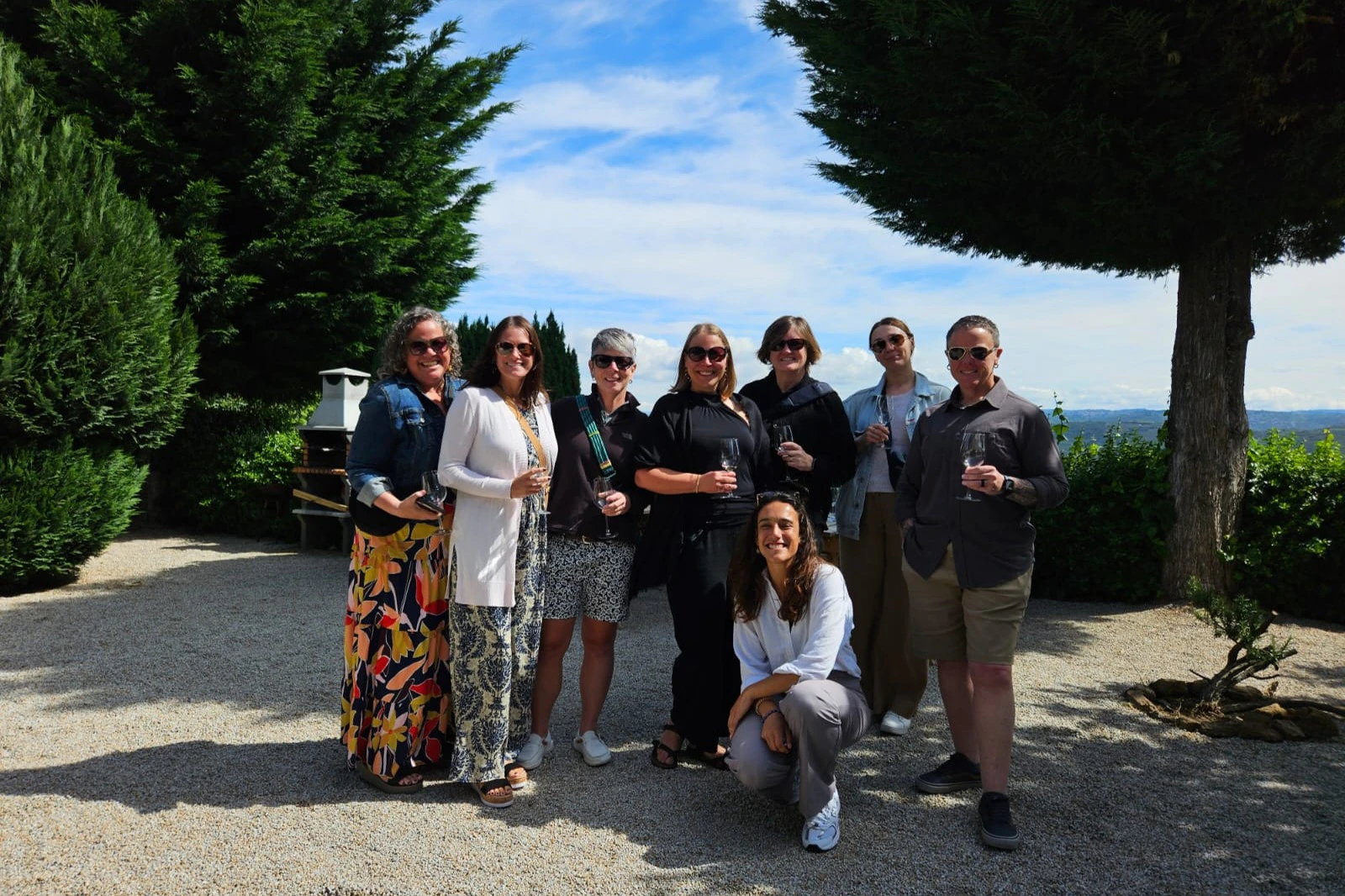 Eight people standing outdoors, smiling, and holding drinks. They are surrounded by greenery under a blue sky with scattered clouds.