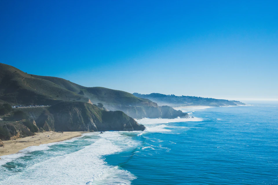 A scenic coastal view showing waves crashing against cliffs and a sandy beach under a clear blue sky.