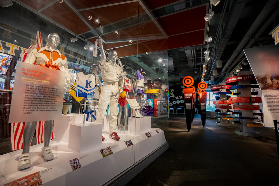 Exhibit featuring cheerleading uniforms and paraphernalia in glass cases, with informative displays and modern signage in the background.