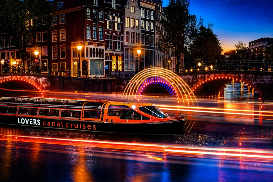 A canal boat with streaks of light trails passes under an illuminated bridge in a city at dusk.