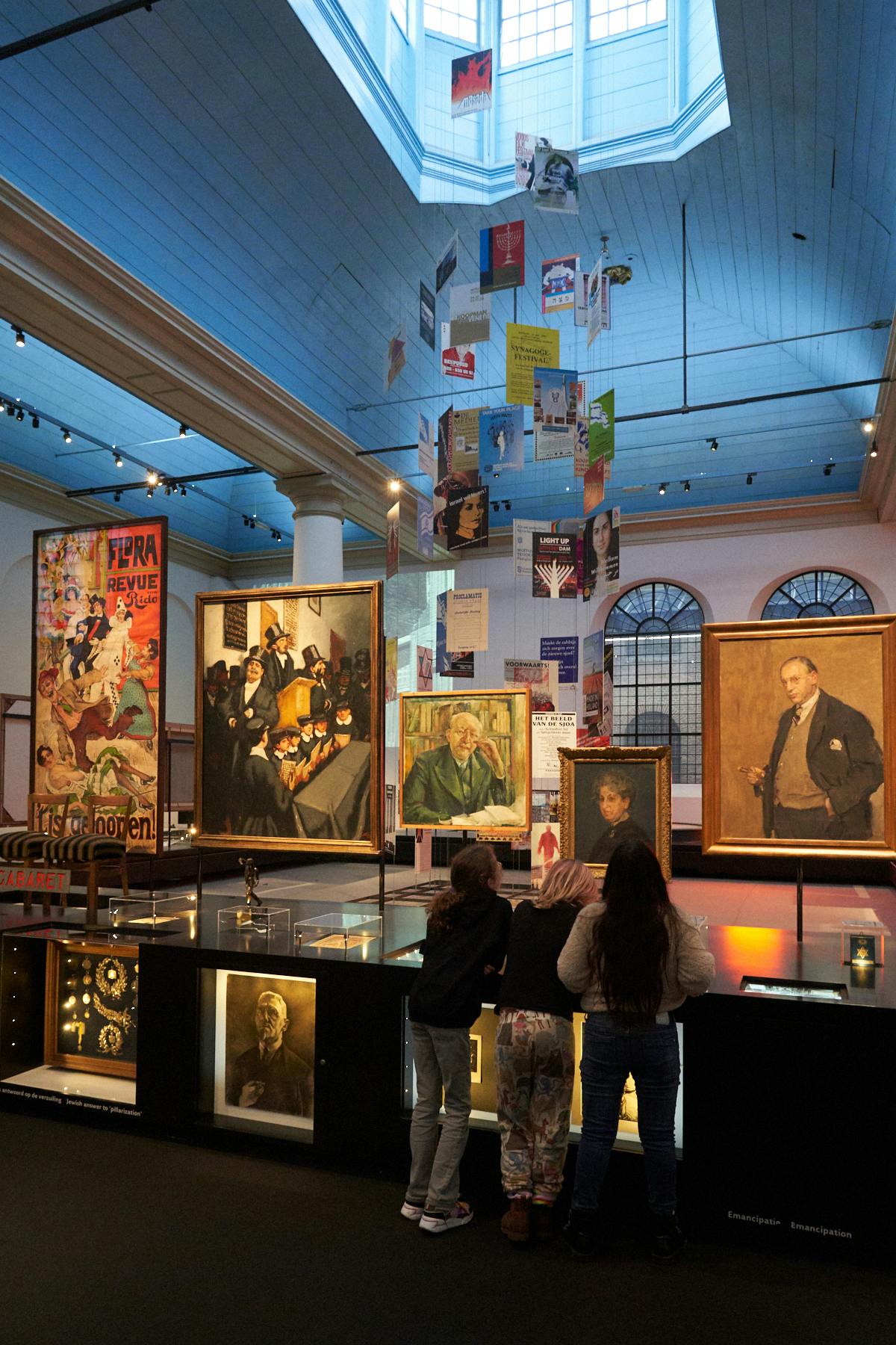 Trois personnes se trouvent dans une galerie d'art où sont exposées des peintures et des affiches encadrées, avec un haut plafond et de grandes fenêtres.