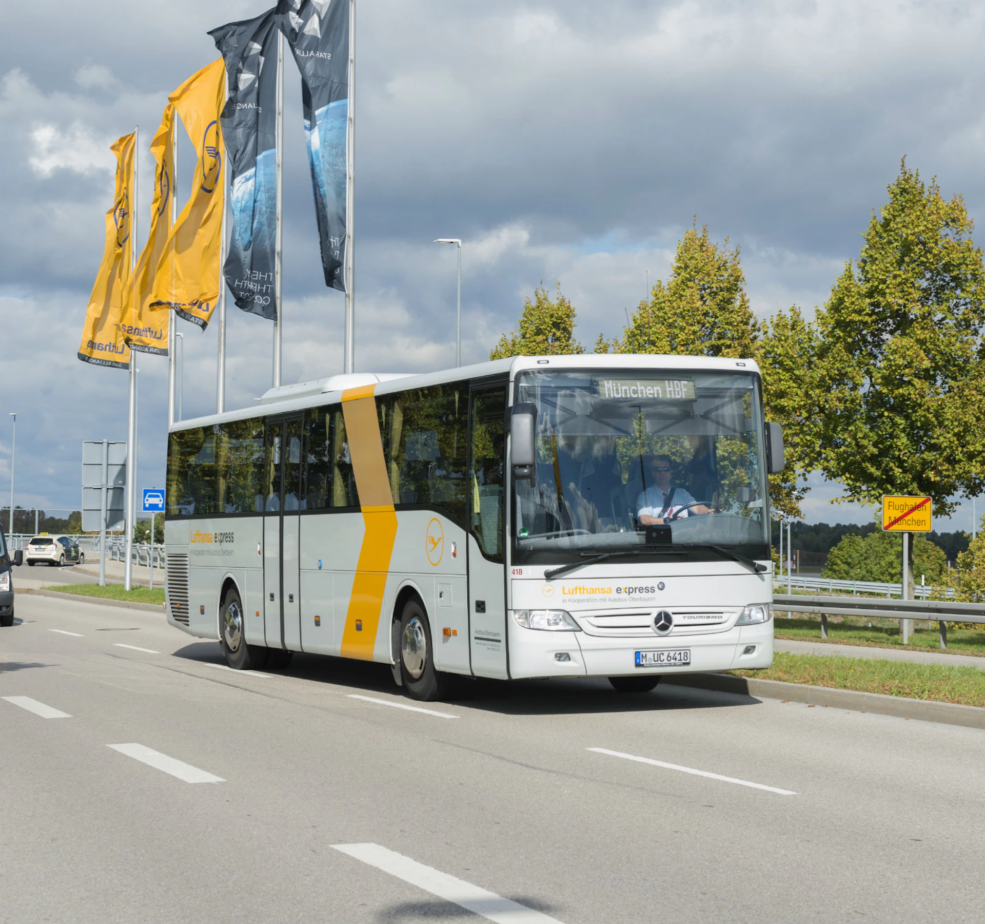 A white Lufthansa Express bus drives on a road with trees and multiple flags in the background.
