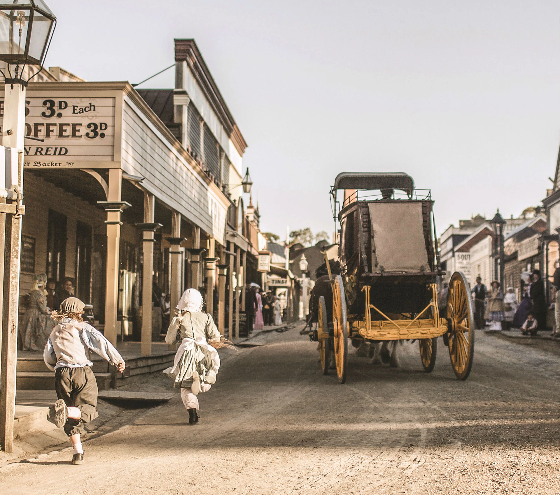 Children running after a horse-drawn carriage on a dirt street in a historic town setting with old-fashioned buildings and signs.