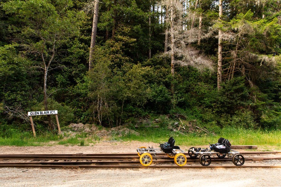 Due carri ferroviari con sedili e pedali sono parcheggiati sui binari del treno di fronte a una foresta rigogliosa. Un cartello recita "R.R. JCT".