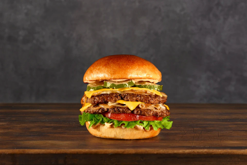 A double cheeseburger with lettuce, tomato, and pickles on a wooden surface against a dark background.