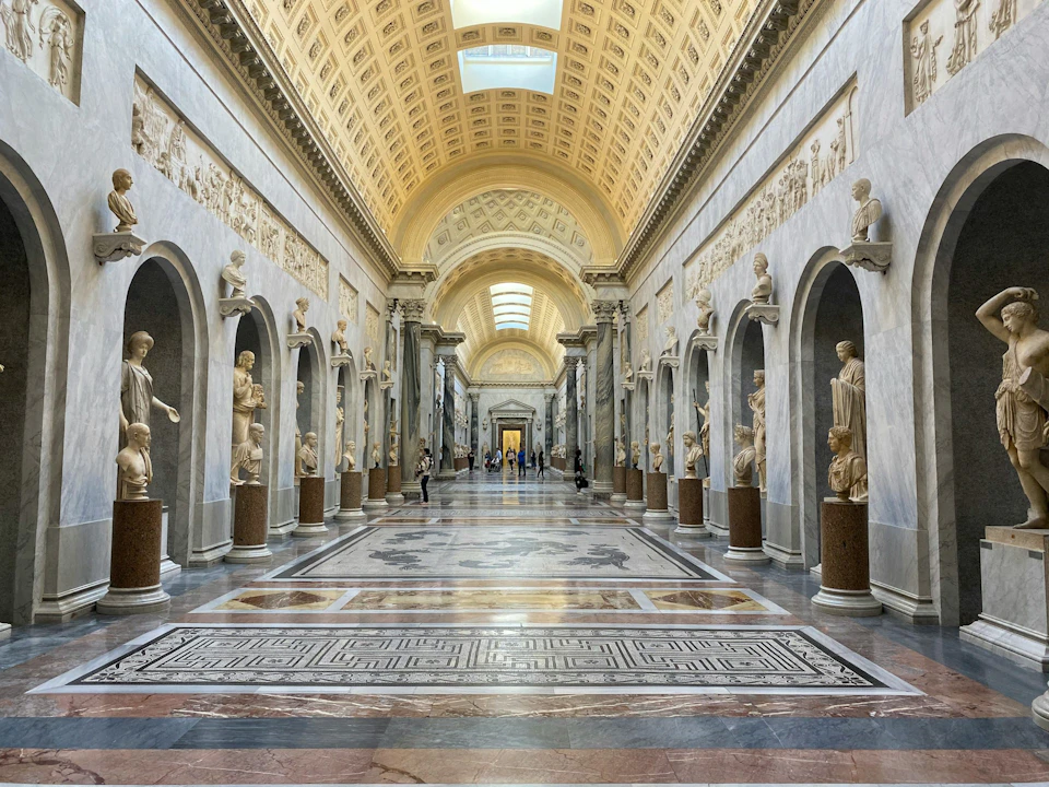 A grand hall with marble statues, intricate carvings, and a vaulted ceiling with skylights. People walk along the patterned floor.
