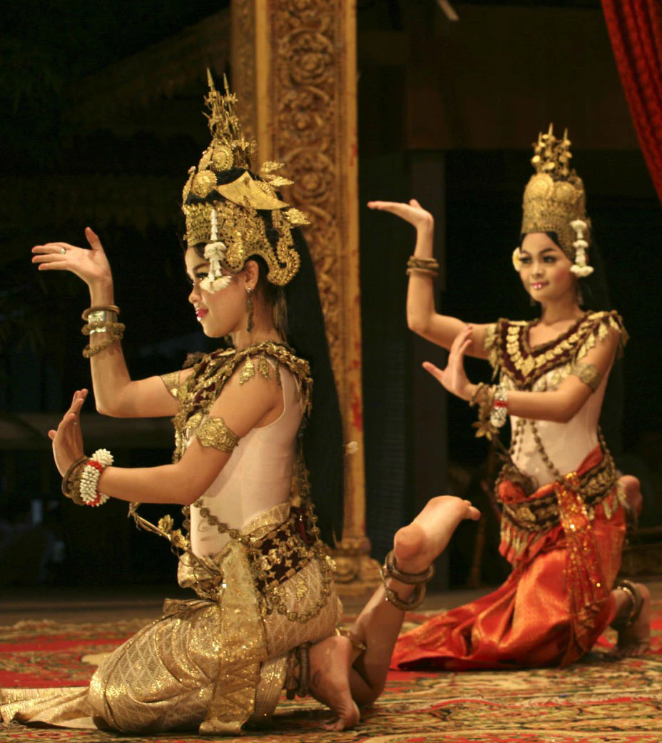 Two dancers in traditional attire perform a classical dance, with ornate headdresses and intricate costumes, in a dimly lit setting.
