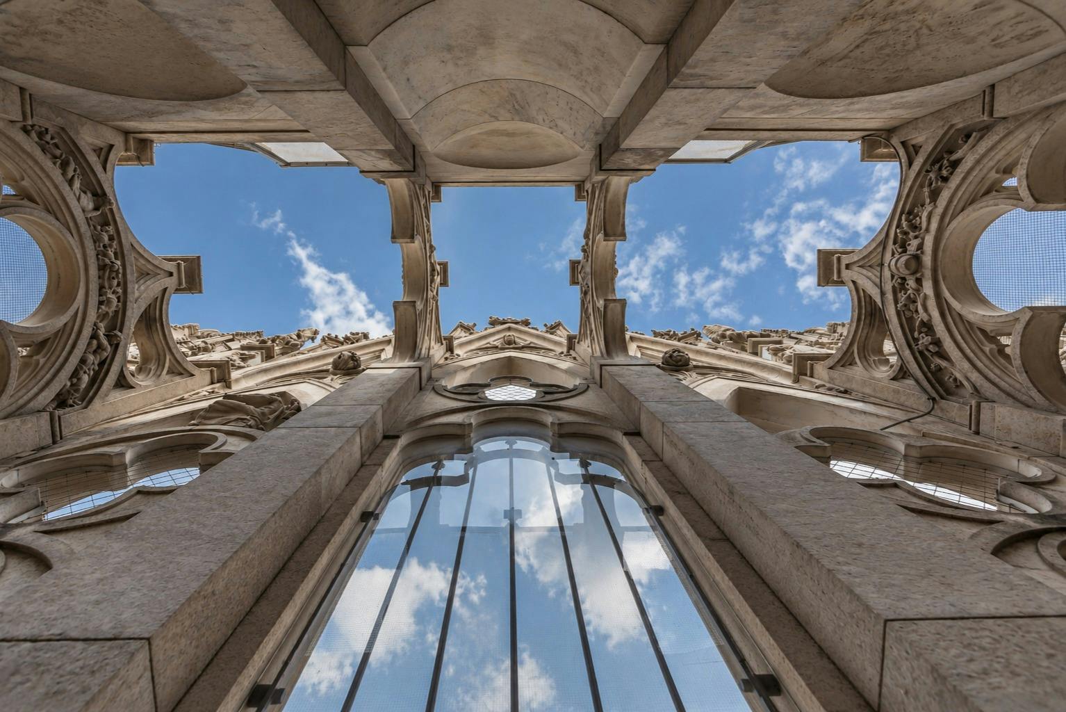 Pohled vzhůru přes zdobnou kamennou architektonickou strukturu se skleněnými okny odhalující modrou oblohu s mraky.