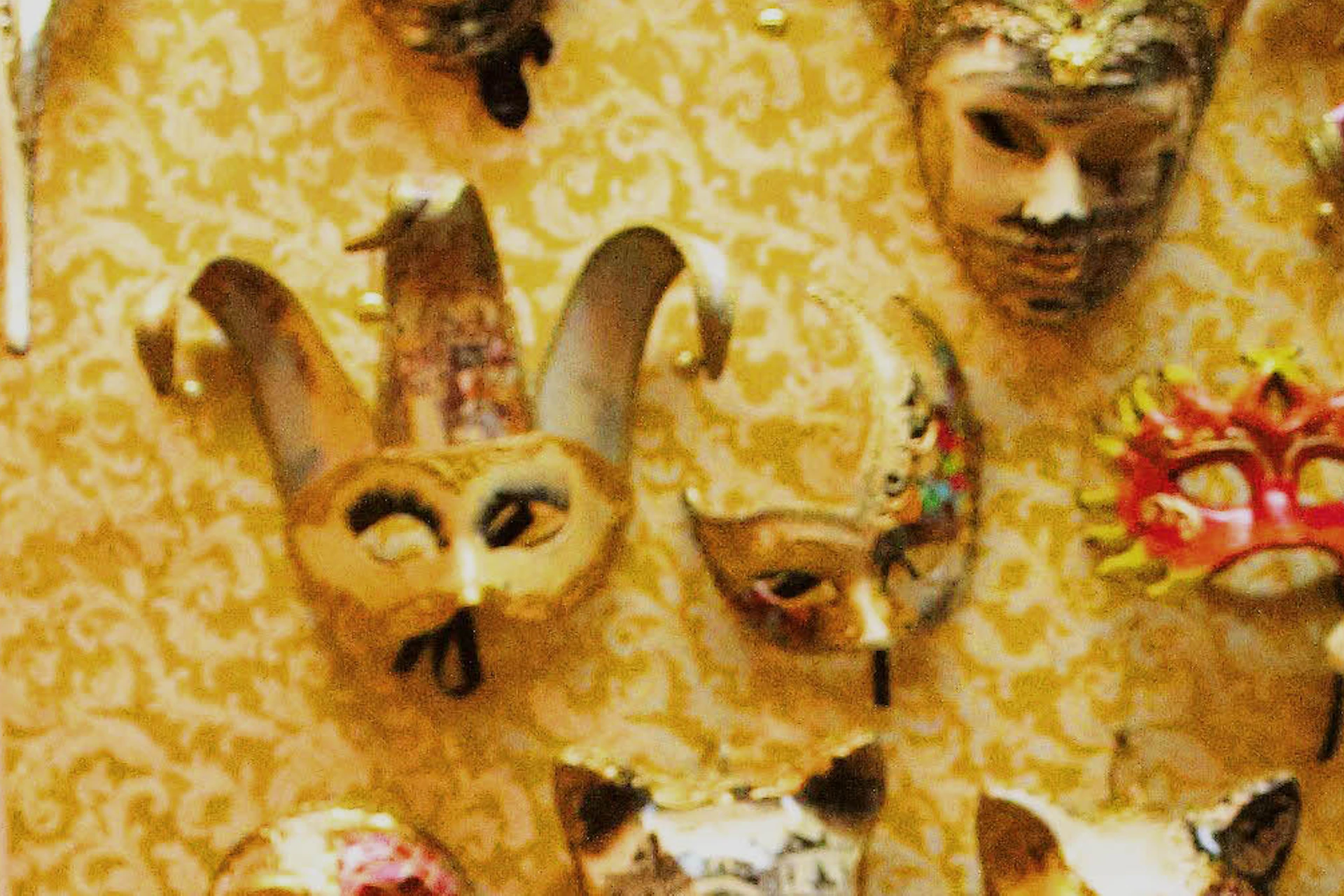 Two people sit at a cluttered table, painting masks in a workshop decorated with various Venetian masks on the wall.