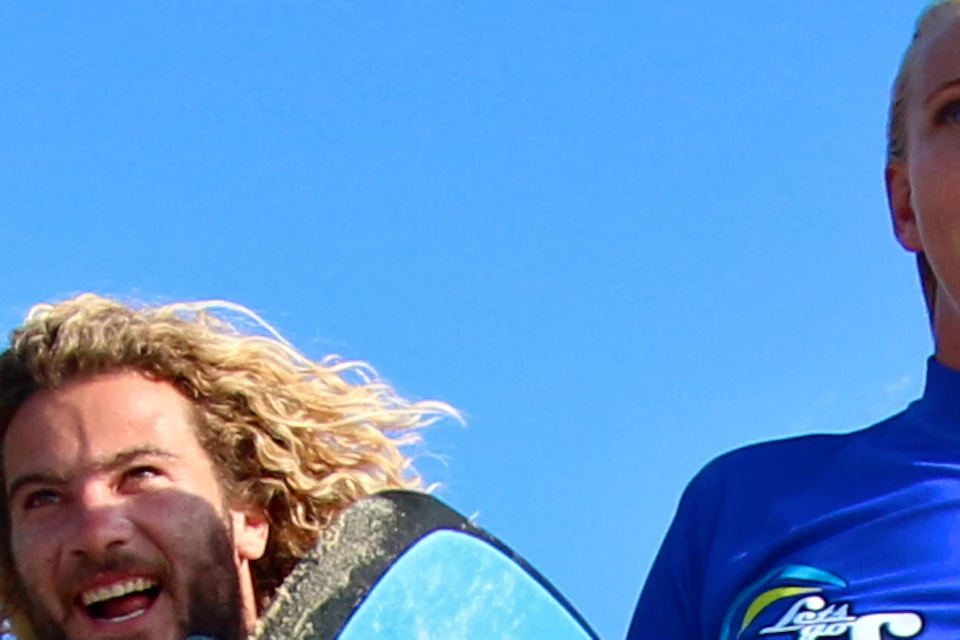 Three people in blue shirts walk barefoot on a beach. One holds a surfboard. Trees and a blue sky are in the background.