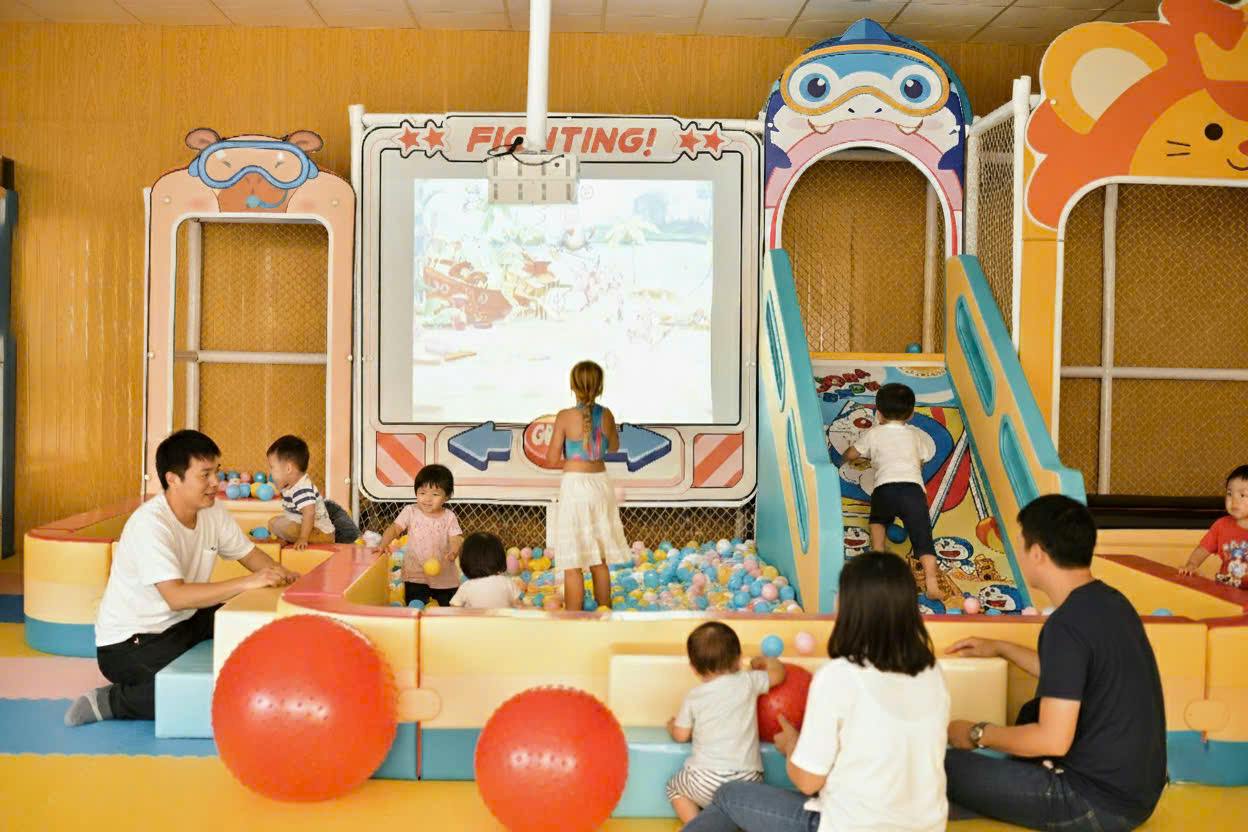 Children and an adult playing in a colorful indoor play area with a ball pit, toys, and a screen displaying a cartoon.