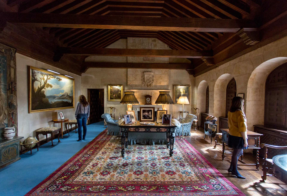 A furnished room with a colorful rug, sofas, table with framed pictures, wall paintings, and two people standing.