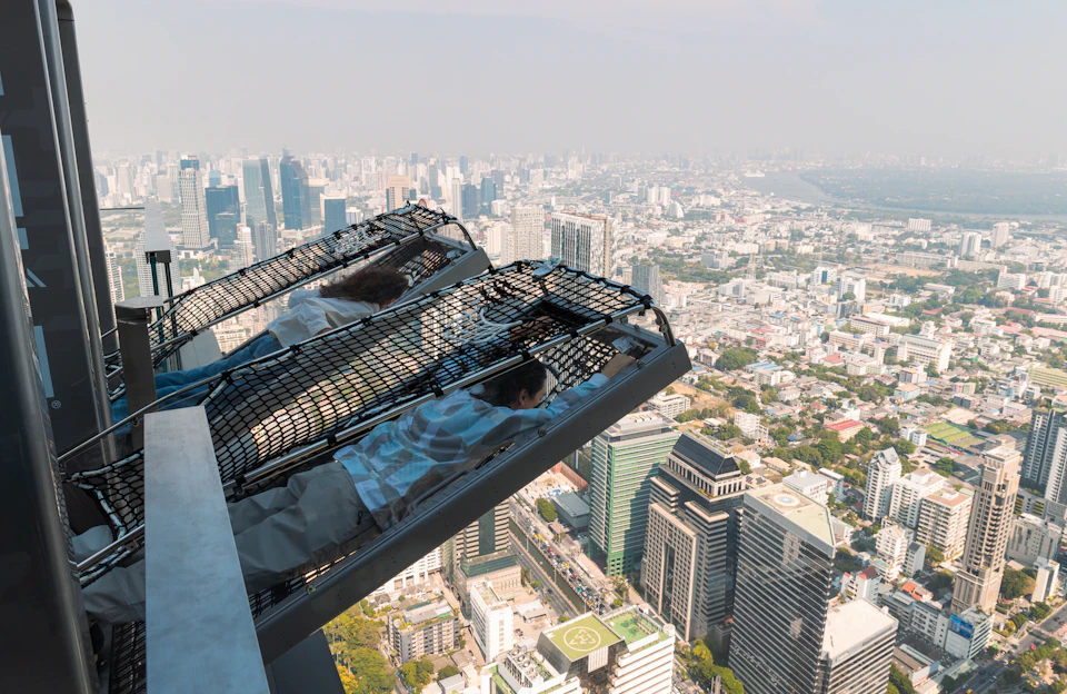 Two people lying face down on an angled, netted platform atop a tall building, overlooking a sprawling cityscape below.
