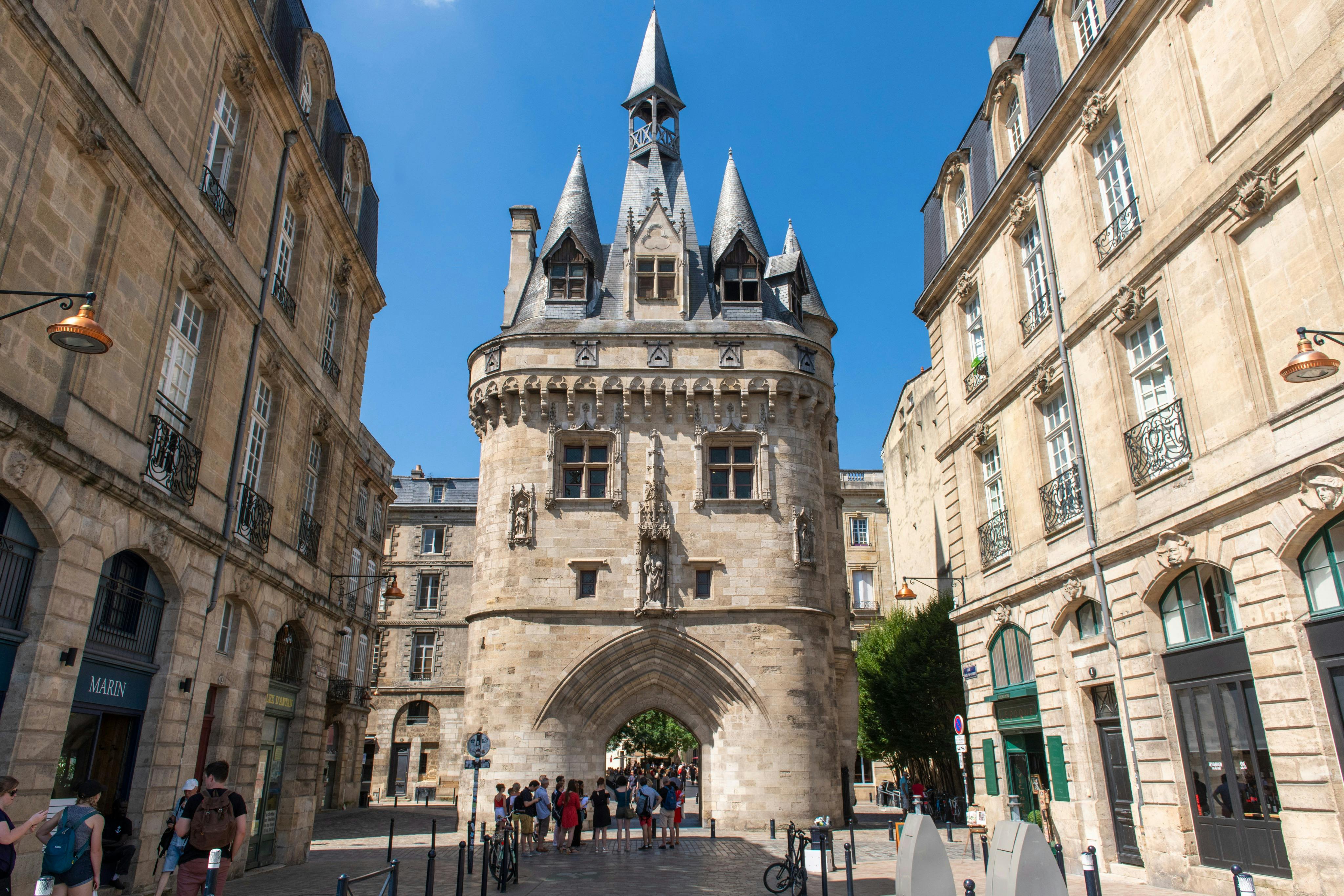 Historical stone gateway with pointed towers and an archway, surrounded by people and old buildings under a clear, blue sky.