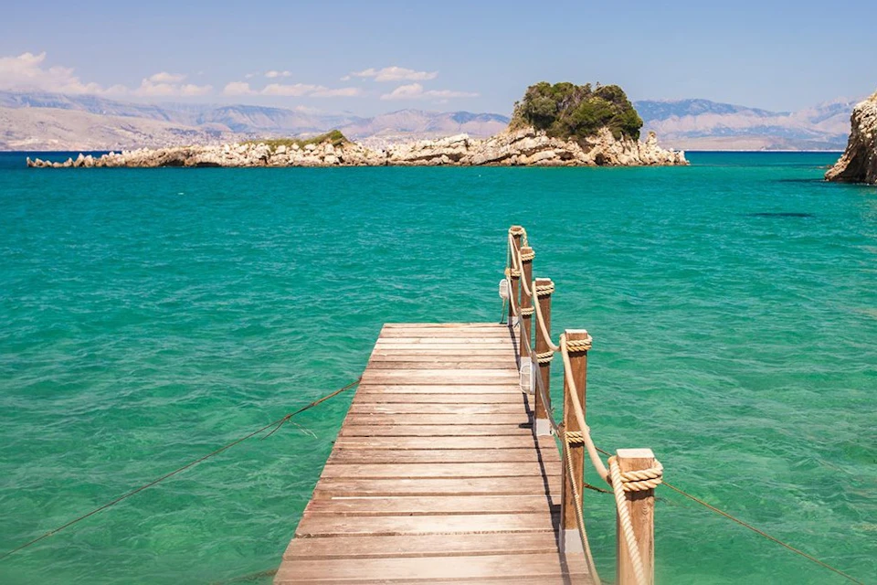 A wooden pier with rope railings extends over turquoise water towards a small rocky island under a clear blue sky.