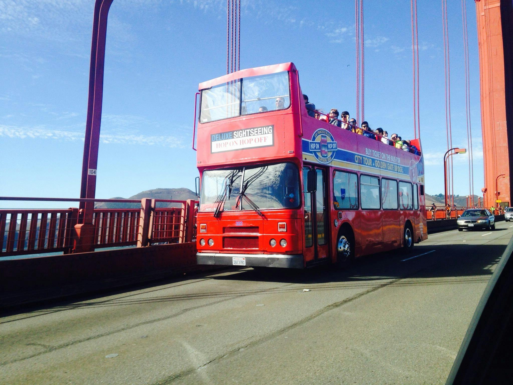 Un bus touristique rouge à impériale avec des passagers traverse un grand pont sous un ciel bleu clair.