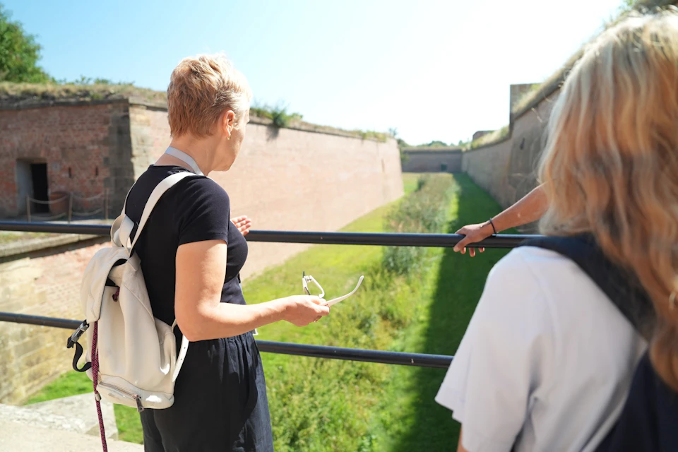 Two people stand near a railing overlooking a grassy trench surrounded by brick walls on a sunny day.