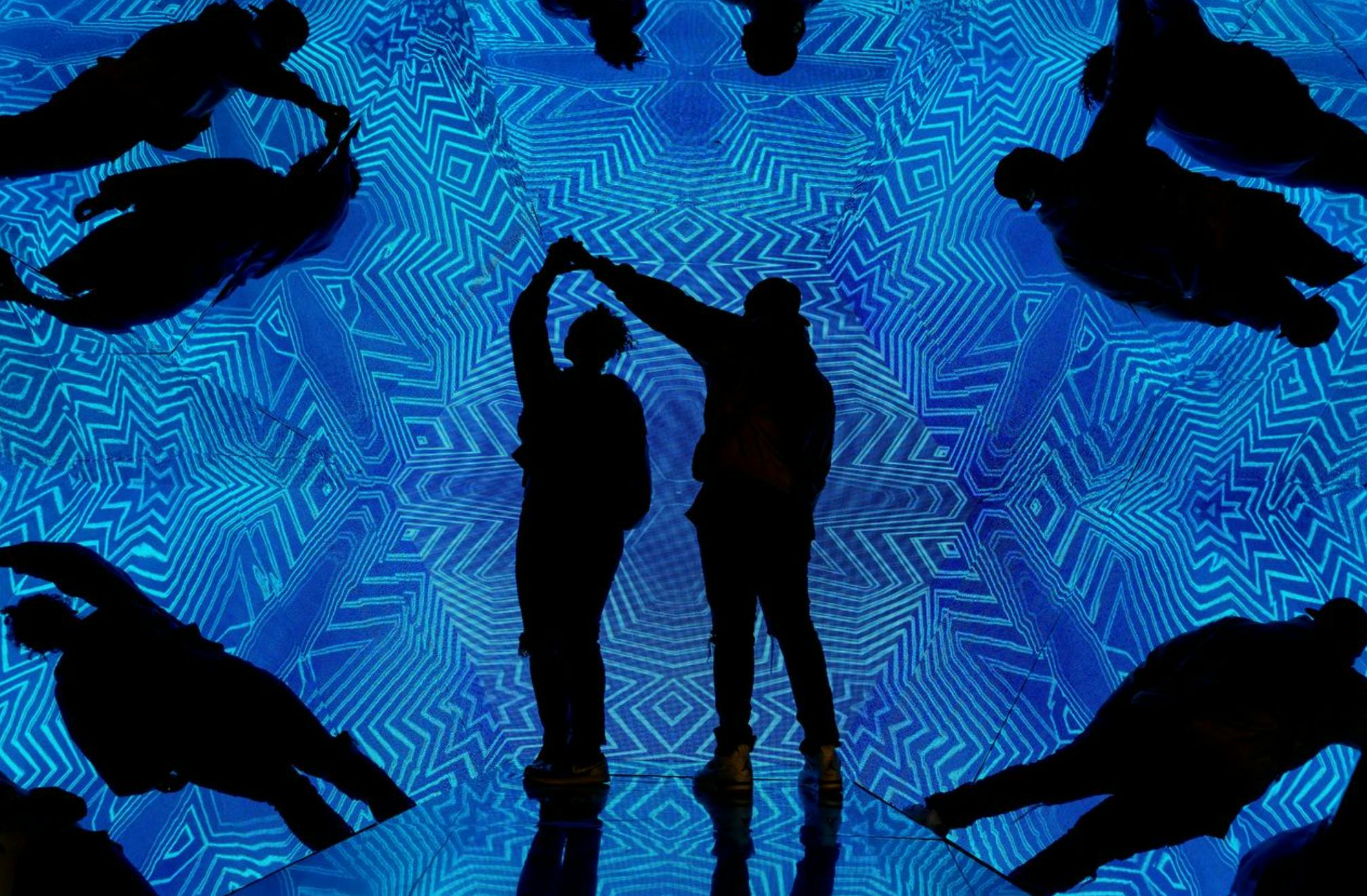 Zwei silhouettierte Figuren tanzen vor einem lebhaften, geometrischen blauen digitalen Hintergrund mit Spiegelungen auf dem Boden.