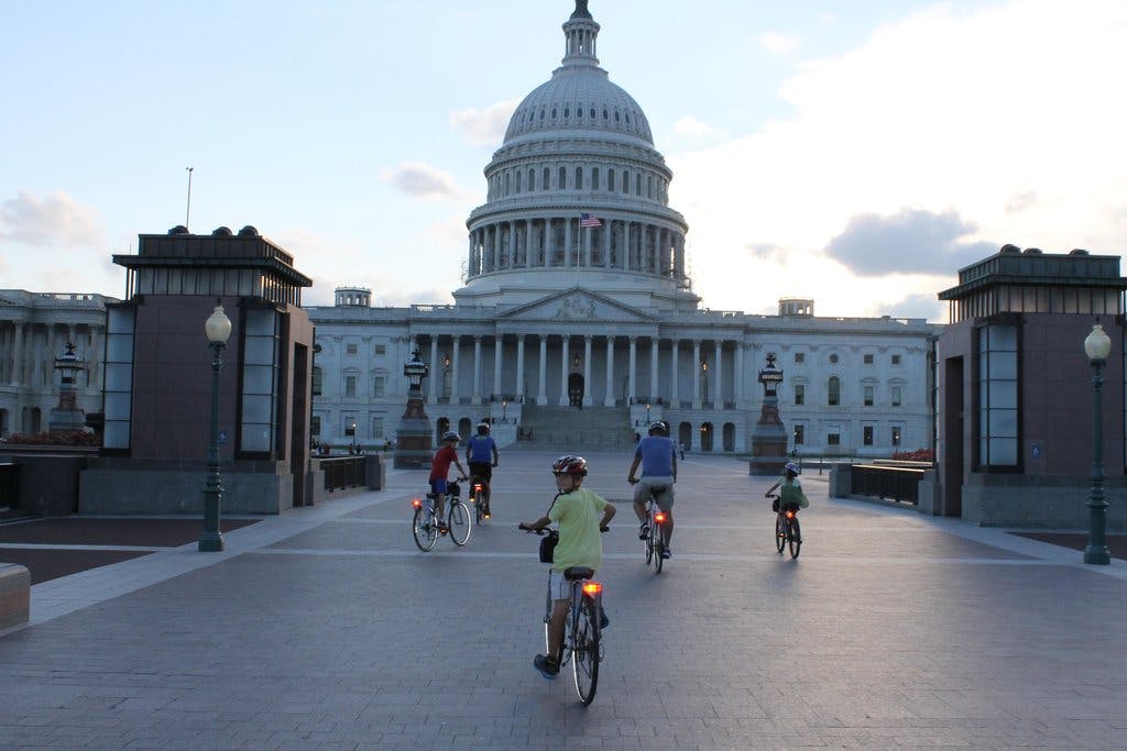 People riding bicycles toward the U.S. Capitol Building under a partly cloudy sky.